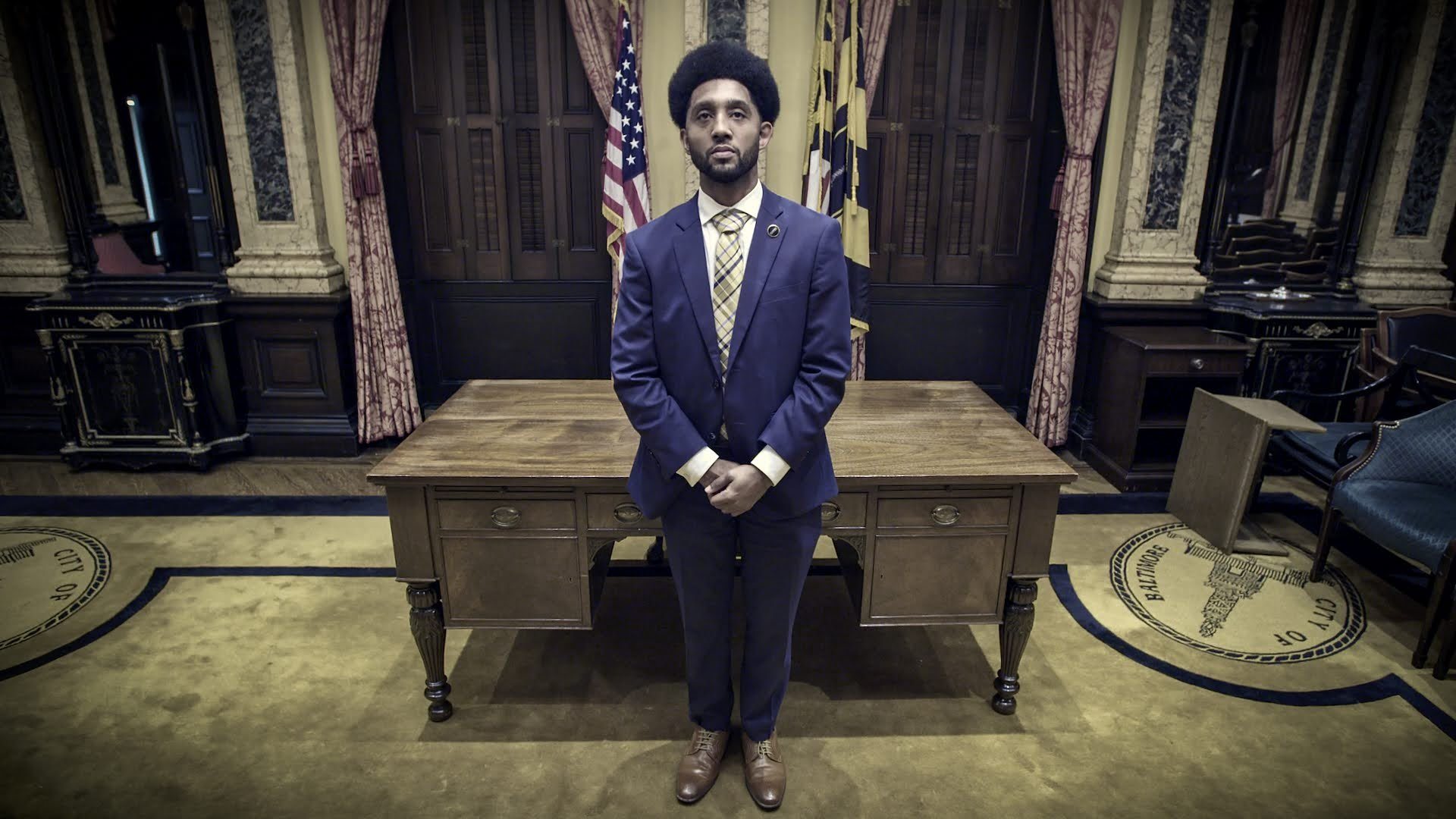 A man in a navy suit stands before Baltimore's City Hall flags and desk — a still from The Body Politic.