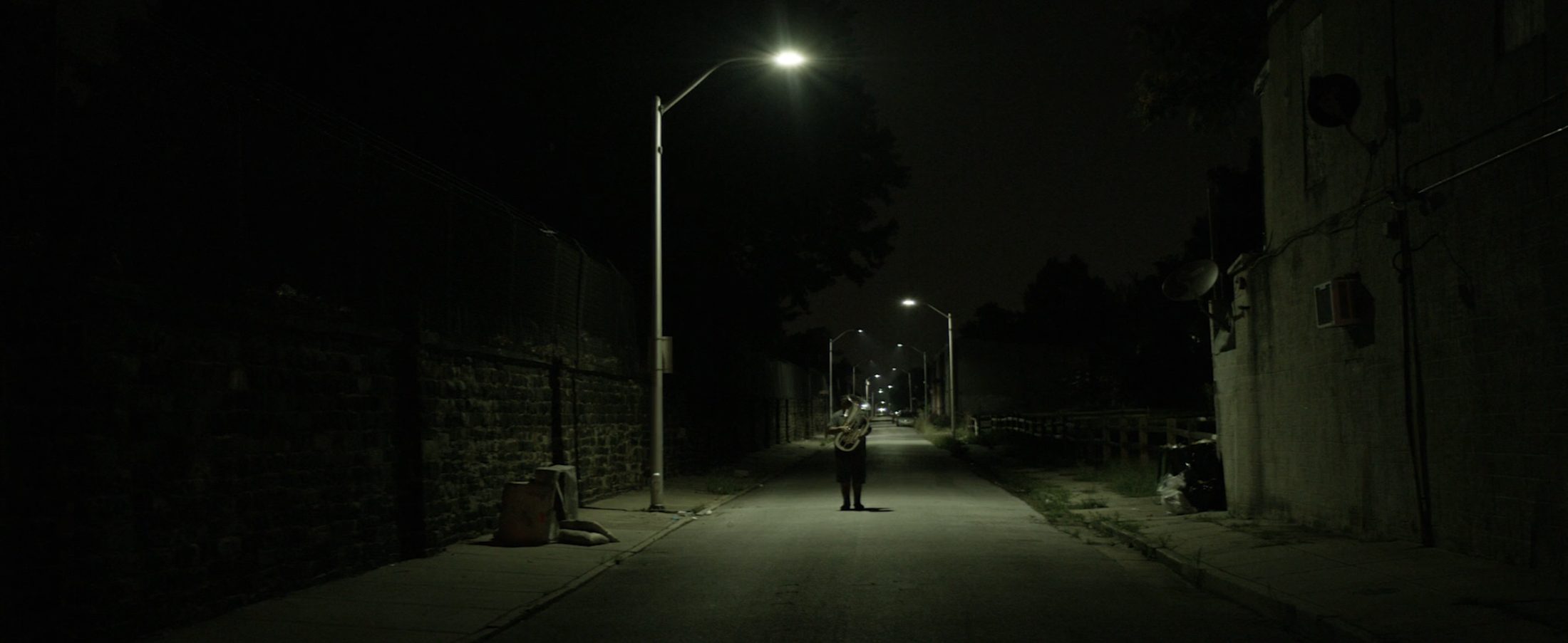 A figure holds a tuba beneath a streetlight on an empty road at night — a still from Hi, I'm Richard Antoine White.