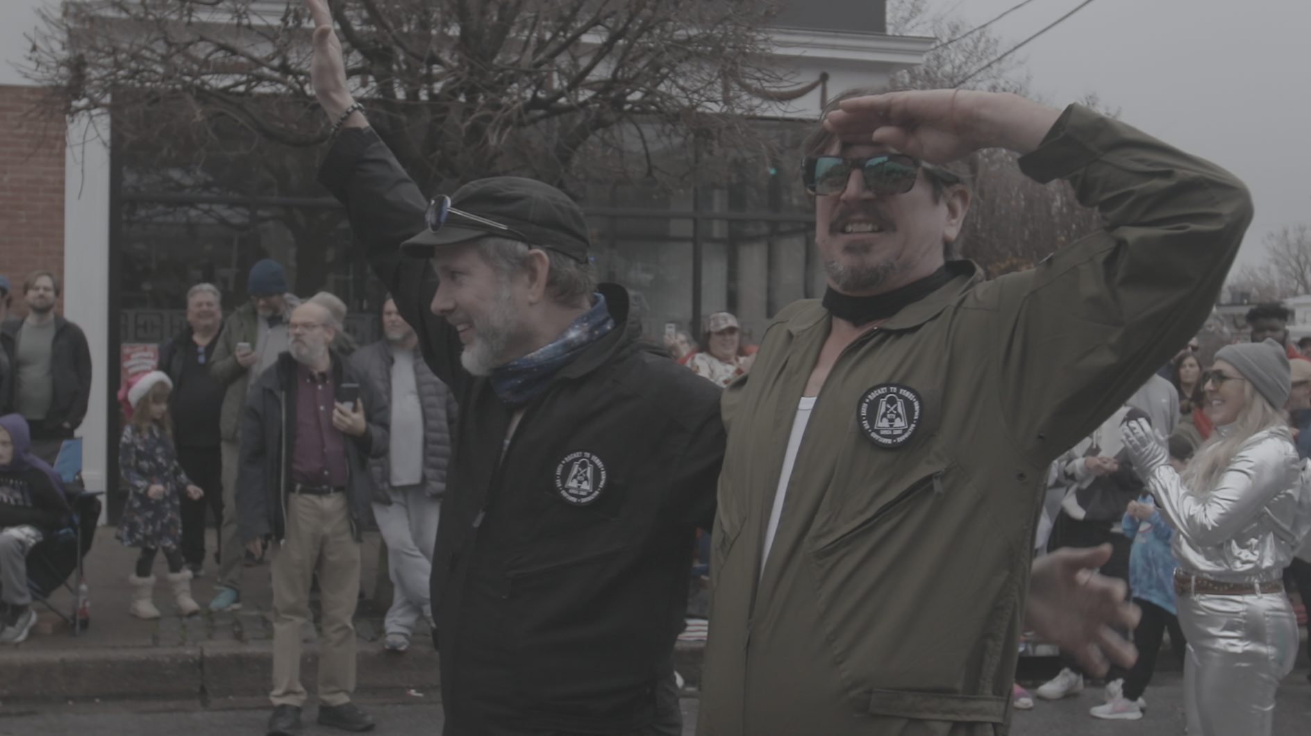 Two men wave to a crowd at an outdoor community event — a still from Rocket to Venus.