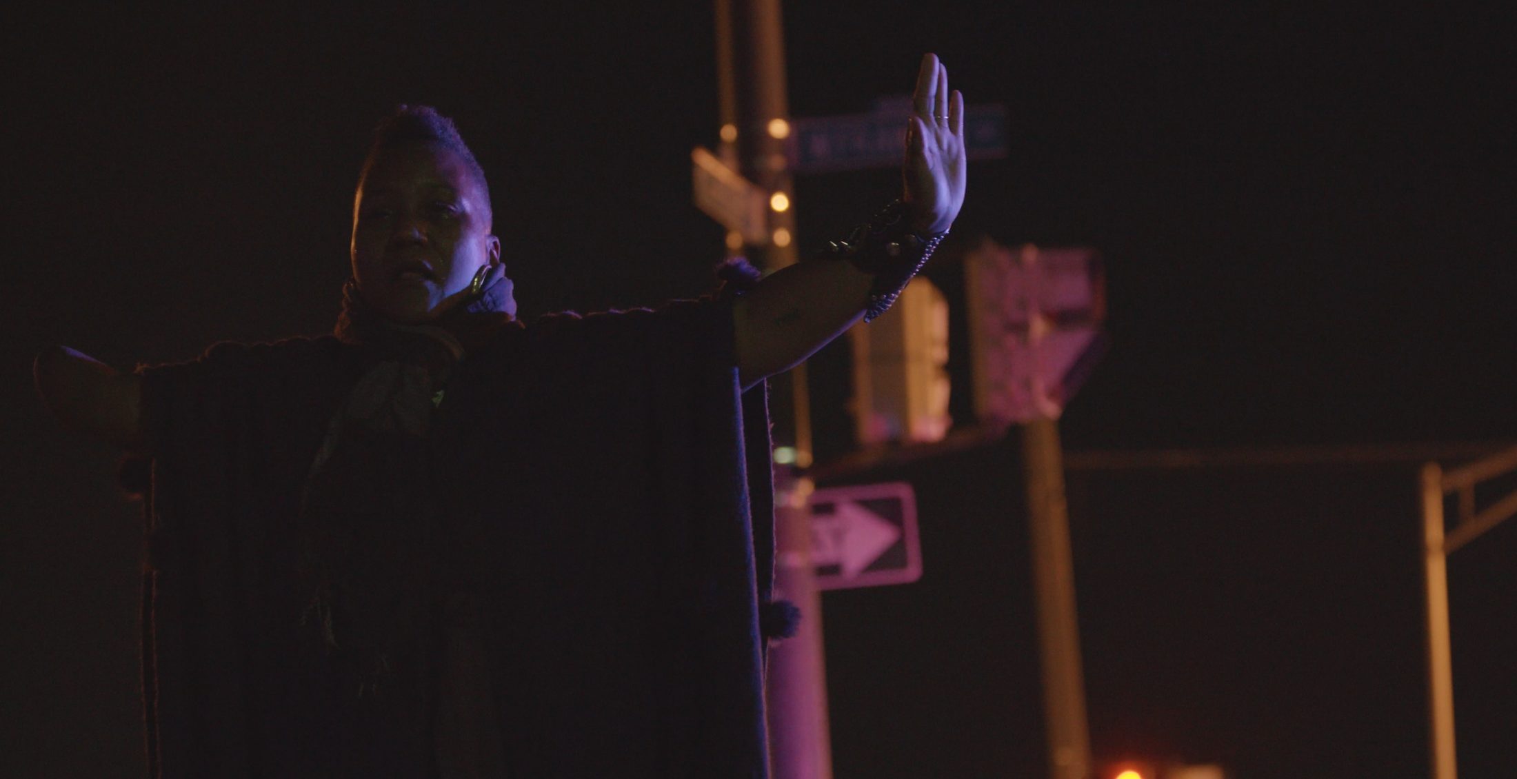 A figure raises both arms beneath streetlights at night, lit in purple and amber — a still from Sage.