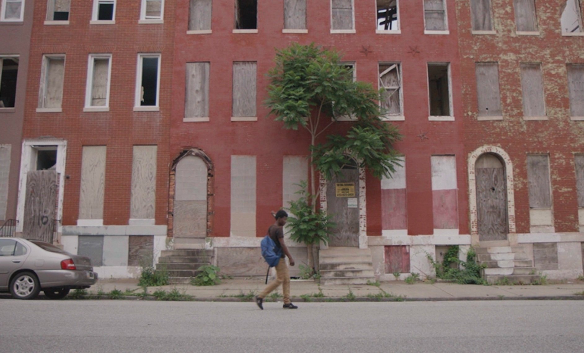 A boy with a backpack walks past a row of boarded-up rowhouses — a still from Squeegee.